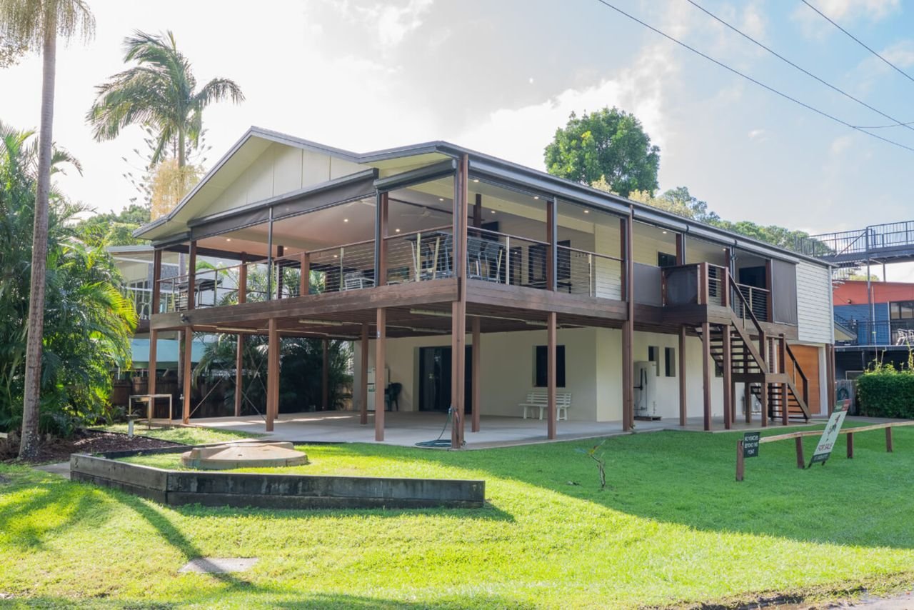 A modern elevated house with a spacious wooden deck, surrounded by greenery and palm trees.